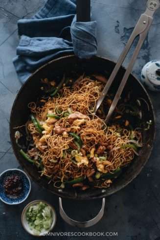 Stir fried thin noodles, chunks of meat, and green vegetables are in a black wok on a blue tablecloth. Green onion garnish sits beside the wok.