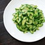 Soft green and yellow slices of cooked cucumbers are piled in a while bowl with a black background.