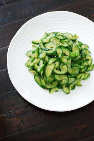 Soft green and yellow slices of cooked cucumbers are piled in a while bowl with a black background.