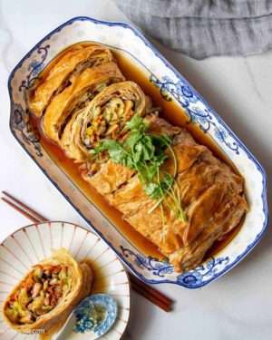 On a blue and white rectangular plate sits a meaty-looking golden sliced roll of tofu skin and vegetables, garnished with a sprig of herbs.
